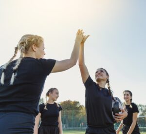 Vier junge Fußballerinnen in schwarzen Trikots geben sich nach einem Spiel oder Training ein High Five auf dem Sportplatz.